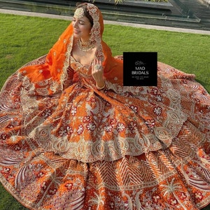 Puede incluir: Un vestido de novia naranja con intrincados bordados y adornos. El vestido presenta una falda en capas y un velo a juego. El vestido est&aacute; adornado con patrones florales blancos y rojos y detalles dorados. La imagen incluye el texto "MAD BRIDALS".