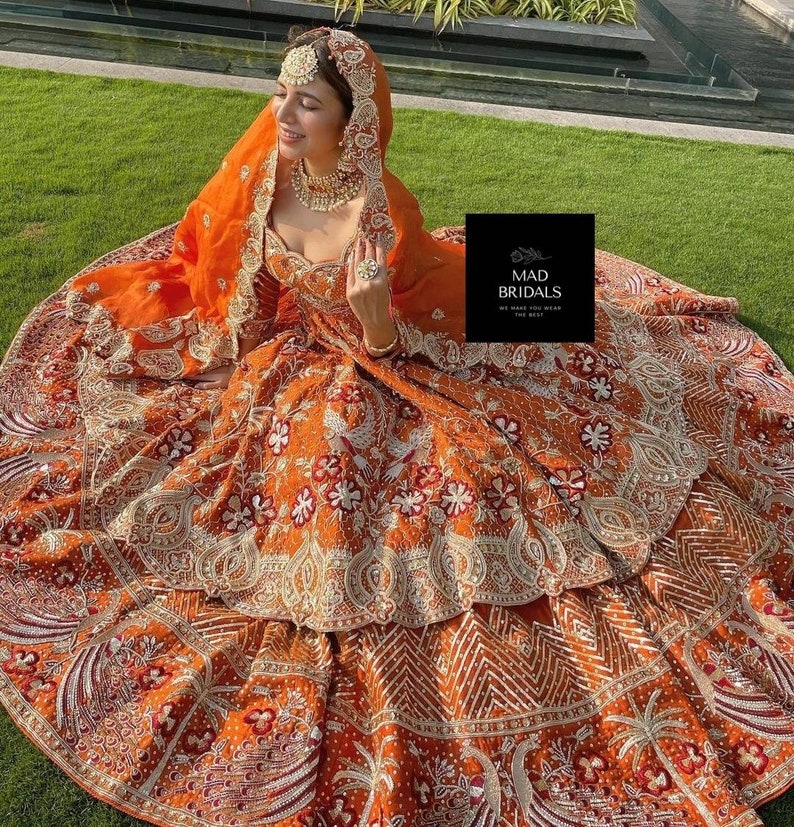 Puede incluir: Un vestido de novia naranja con intrincados bordados y adornos. El vestido presenta una falda en capas y un velo a juego. El vestido est&aacute; adornado con patrones florales blancos y rojos y detalles dorados. La imagen incluye el texto "MAD BRIDALS".
