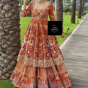 Puede incluir: Un vestido largo naranja con intrincados bordados dorados y una falda escalonada. El vestido presenta mangas largas y un escote cuadrado. La imagen incluye el texto "MAD BRIDALS".
