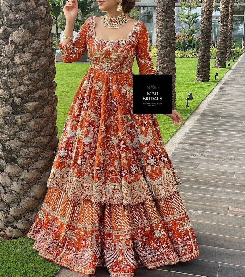 Puede incluir: Un vestido largo naranja con intrincados bordados dorados y una falda escalonada. El vestido presenta mangas largas y un escote cuadrado. La imagen incluye el texto "MAD BRIDALS".