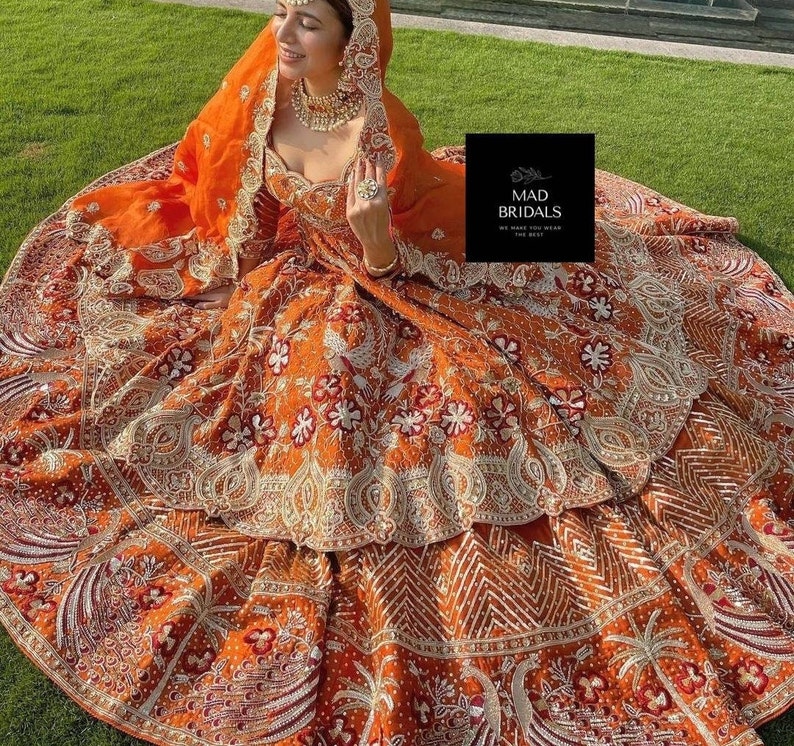 Puede incluir: Un vestido de novia naranja con intrincados bordados y adornos. El vestido presenta una falda detallada con patrones florales y geom&eacute;tricos, un velo a juego y un corpi&ntilde;o con escote coraz&oacute;n. La imagen incluye el texto "MAD BRIDALS".