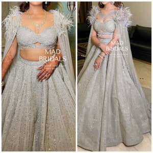 Puede incluir: Un conjunto lehenga plateado que presenta un top corto muy adornado con escote coraz&oacute;n y una falda larga a juego. El atuendo incluye una capa transparente con adornos de plumas en los hombros. El texto en la imagen dice MAD BRIDALS.
