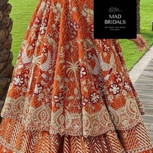 Puede incluir: Un vestido formal bordado de color rojo anaranjado con intrincados patrones florales y de aves dorados y blancos. El vestido presenta una falda escalonada y mangas largas. La imagen incluye el texto "MAD BRIDALS WE MAKE YOU WEAR THE BEST".