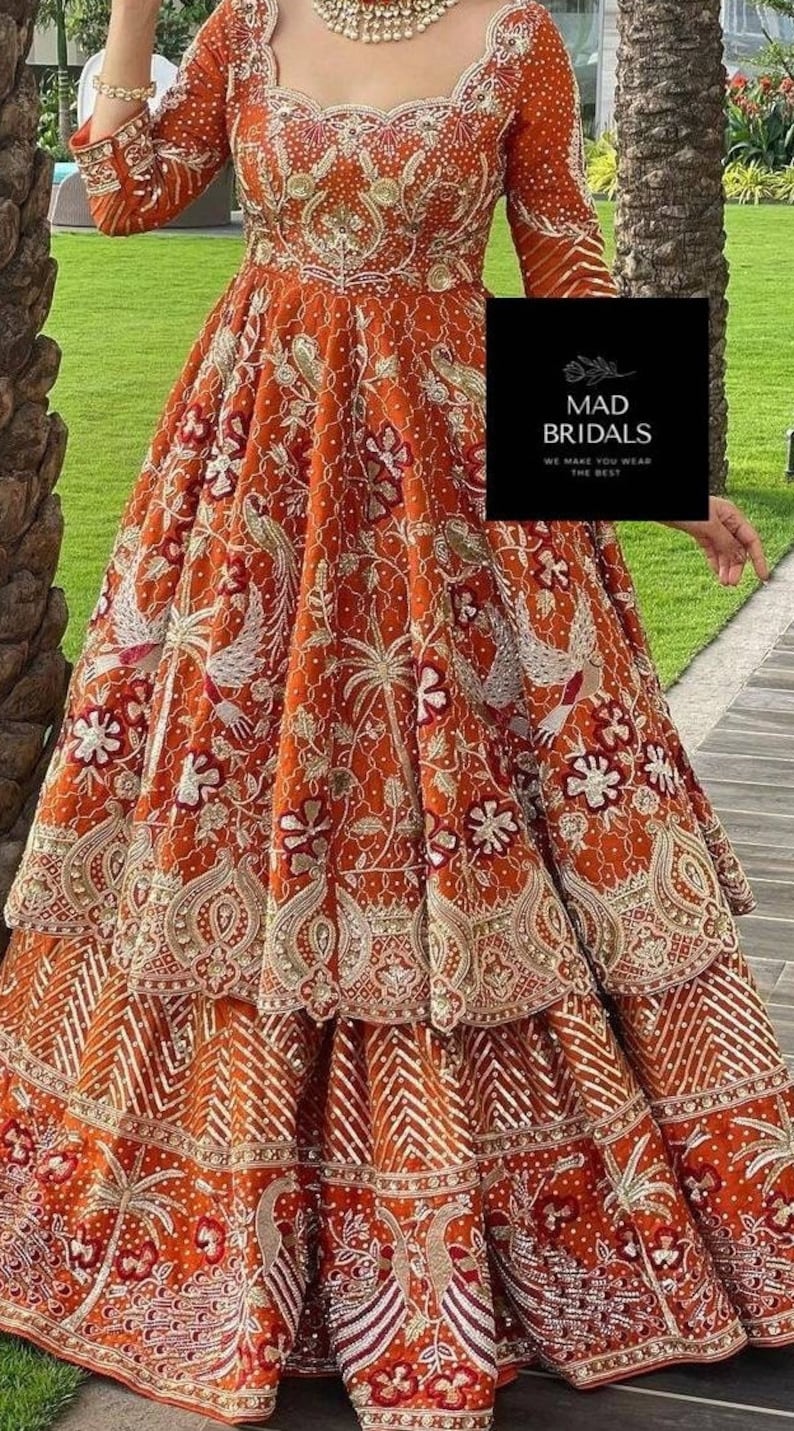 Puede incluir: Un vestido formal bordado de color rojo anaranjado con intrincados patrones florales y de aves dorados y blancos. El vestido presenta una falda escalonada y mangas largas. La imagen incluye el texto "MAD BRIDALS WE MAKE YOU WEAR THE BEST".