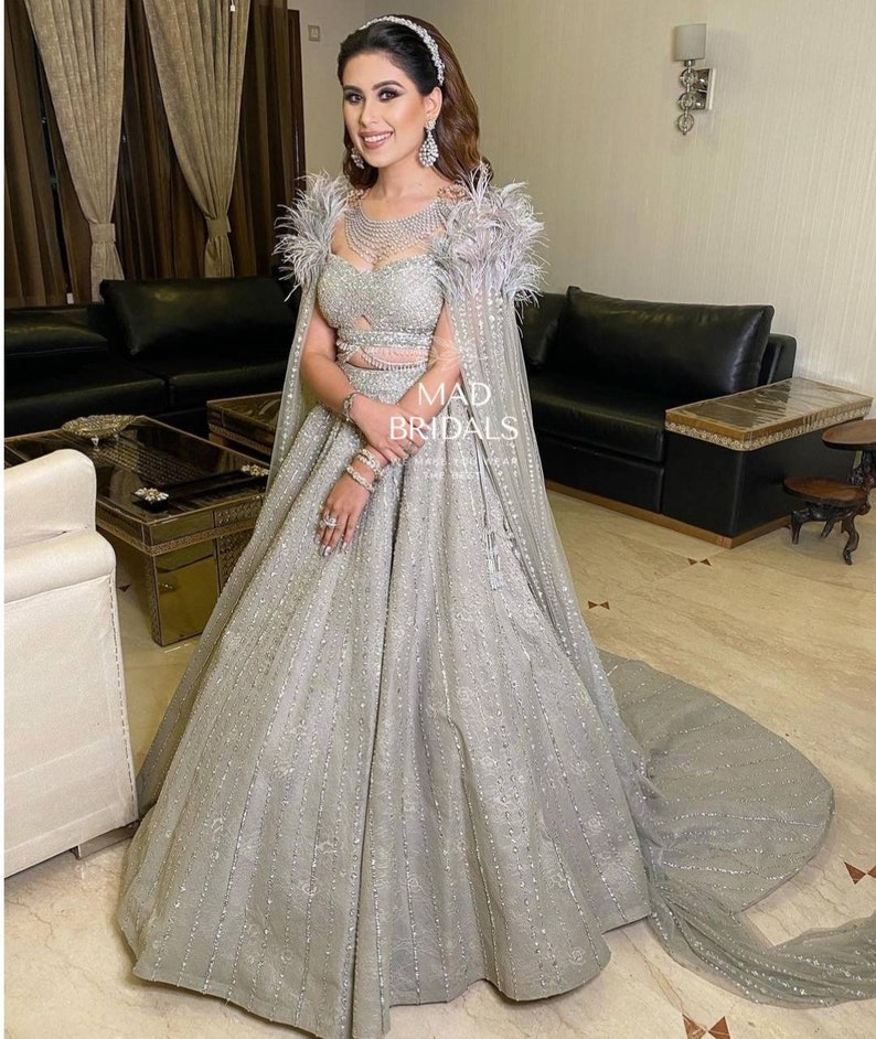 Puede incluir: Un vestido plateado hasta el suelo con escote coraz&oacute;n, abdomen descubierto y una capa a juego con hombros adornados con plumas. La prenda est&aacute; muy adornada con lentejuelas y bordados plateados, incluyendo el texto MAD BRIDALS.