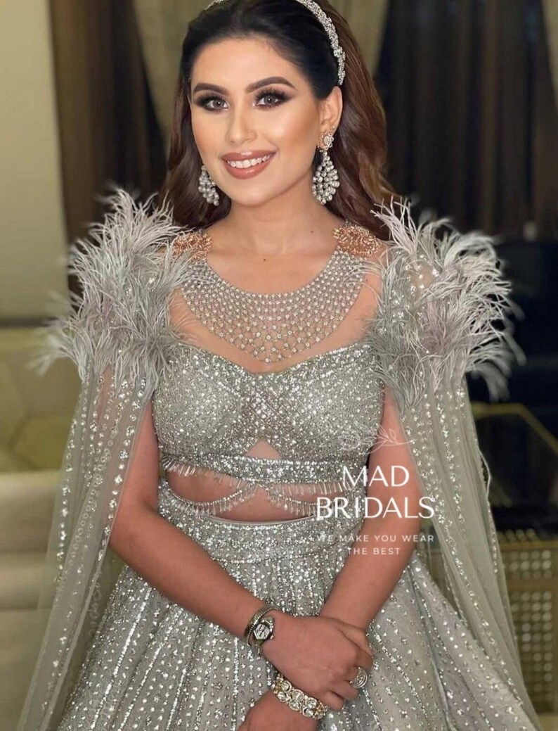 Puede incluir: Un conjunto de dos piezas plateado que presenta un corpi&ntilde;o corto y una falda de talle alto, ambos muy adornados con lentejuelas y cuentas. El atuendo incluye una capa transparente con hombros adornados con plumas y el texto MAD BRIDALS WE MAKE YOU WEAR THE BEST.