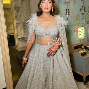 Puede incluir: Un conjunto lehenga plateado que presenta un top corto muy adornado con hombros decorados con plumas y una falda larga a juego con patrones de lentejuelas verticales. El atuendo incluye una capa transparente y lleva el texto MAD BRIDALS.