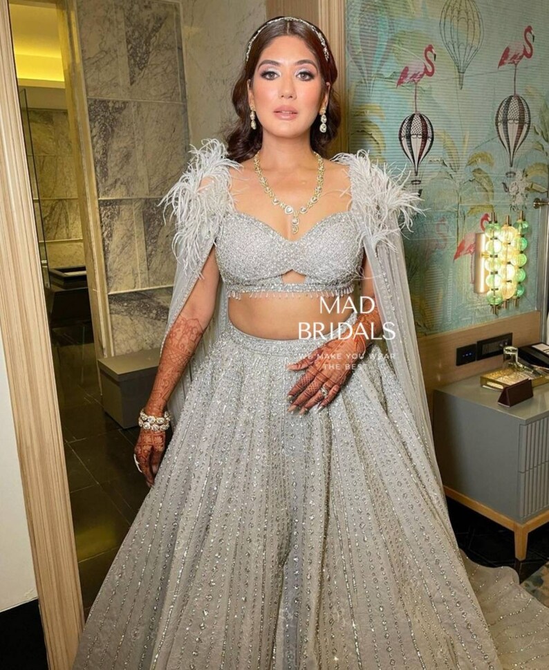 Puede incluir: Un conjunto lehenga plateado que presenta un top corto muy adornado con hombros decorados con plumas y una falda larga a juego con patrones de lentejuelas verticales. El atuendo incluye una capa transparente y lleva el texto MAD BRIDALS.