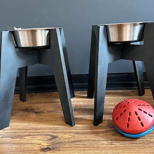 Könnte beinhalten: Zwei schwarze erhöhte Hundenäpfe mit Edelstahlschalen oben. Die Näpfe stehen auf einem Holzboden mit einem roten und blauen Hundespielzeug davor.