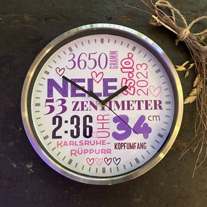 Könnte beinhalten: Eine weiße Uhr mit silbernem Rahmen zeigt ein lila-rosa Design mit dem Text "Nele 3650 Gramm 53 Zentimeter 2:36 Uhr 34 cm Karlsruhe-Ruppurr Kopfumfang".
