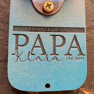 May include: A light blue wooden bottle opener with a brass bottle opener attached. The bottle opener has the text "Der Weltbeste Papa Klara Est. 2019" engraved on it.