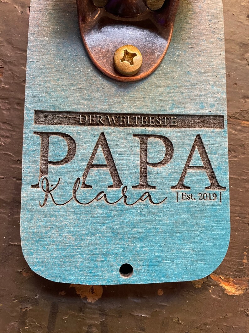 May include: A light blue wooden bottle opener with a brass bottle opener attached. The bottle opener has the text "Der Weltbeste Papa Klara Est. 2019" engraved on it.