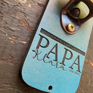 May include: A blue and black wooden bottle opener with a copper bottle opener attached. The bottle opener has the text "DER WELTBESTE PAPA klara | Est. 2019 |" engraved on it.