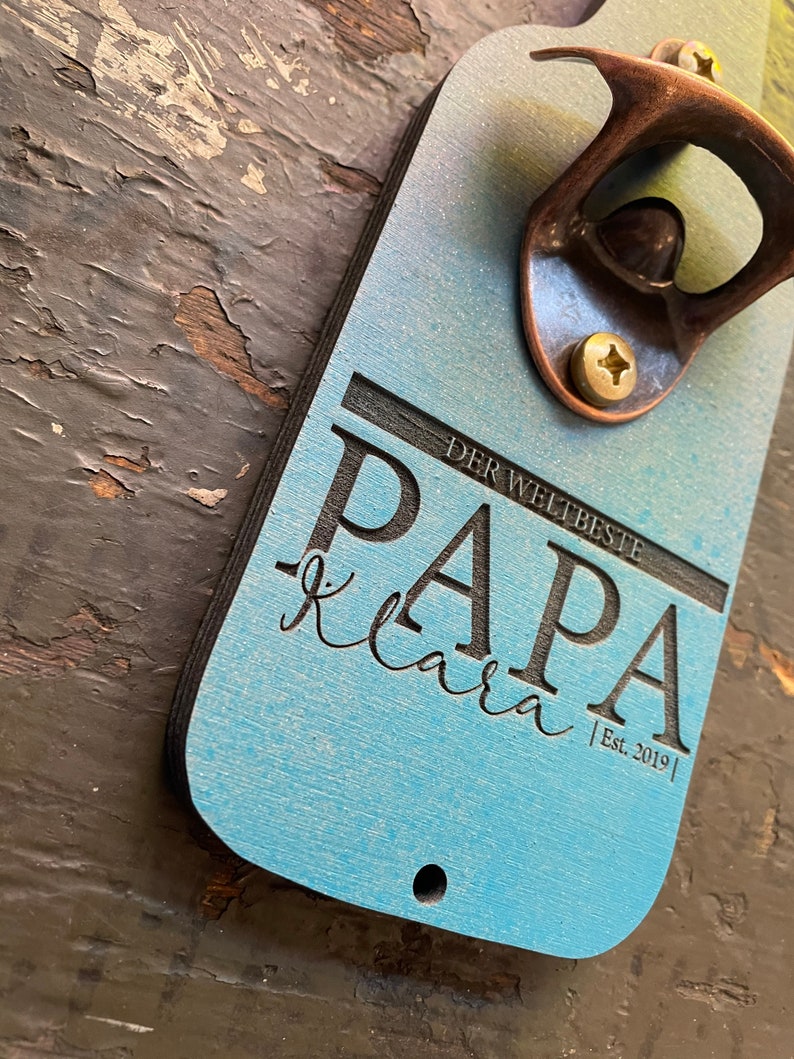 May include: A blue and black wooden bottle opener with a copper bottle opener attached. The bottle opener has the text "DER WELTBESTE PAPA klara | Est. 2019 |" engraved on it.