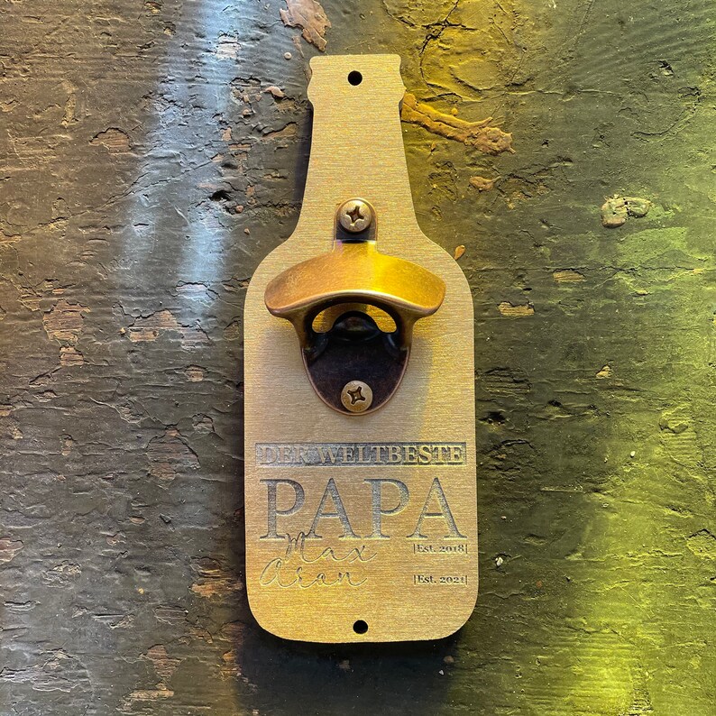 May include: A wooden bottle opener shaped like a beer bottle with a brass bottle opener. The bottle opener has the text "Der Weltbeste Papa" and "Max Allen" with the dates "Est. 2018" and "Est. 2021".
