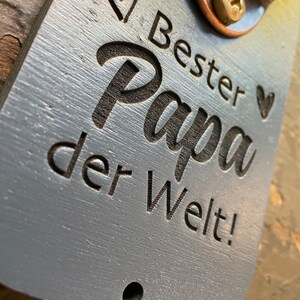 May include: A gray wooden bottle opener with a metal bottle opener attached. The bottle opener has the text "Bester Papa der Welt!" engraved on it.