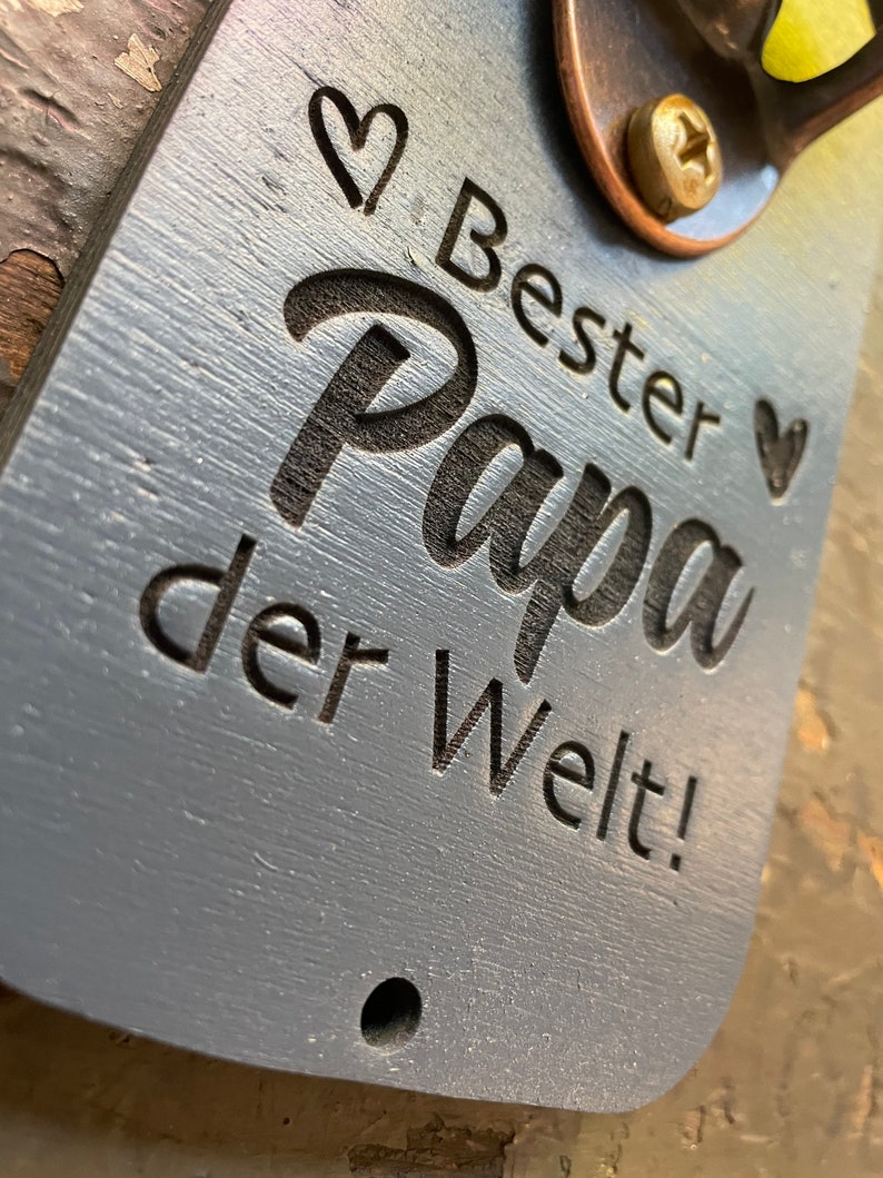 May include: A gray wooden bottle opener with a metal bottle opener attached. The bottle opener has the text "Bester Papa der Welt!" engraved on it.