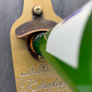 May include: A gold-colored bottle opener with a black screw and a decorative design that says "Ruhplop" for the best Opa in the world.  The bottle opener is mounted on a black slate background.