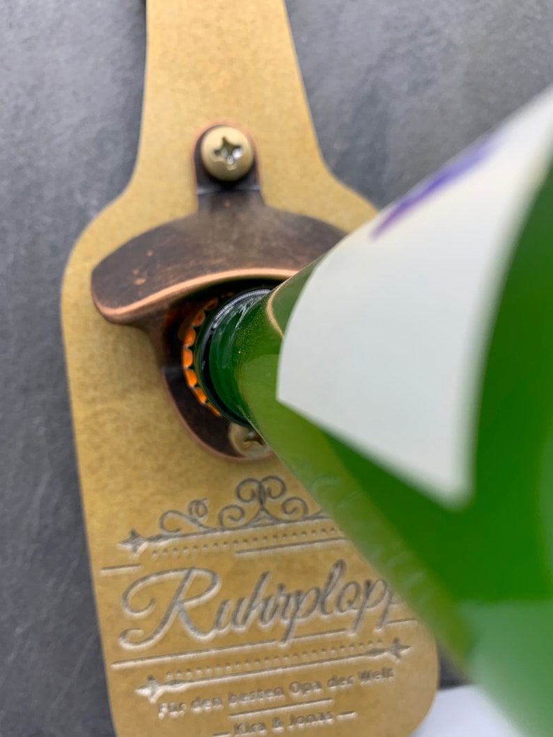May include: A gold-colored bottle opener with a black screw and a decorative design that says "Ruhplop" for the best Opa in the world.  The bottle opener is mounted on a black slate background.