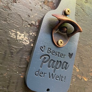 May include: A blue wooden bottle opener shaped like a beer bottle with a copper bottle opener attached. The bottle opener has the text "Bester Papa der Welt!" engraved on it.