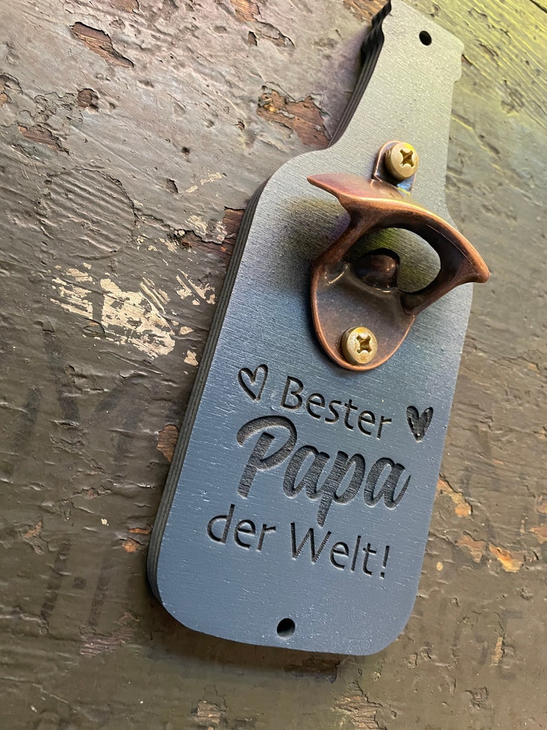 May include: A blue wooden bottle opener shaped like a beer bottle with a copper bottle opener attached. The bottle opener has the text "Bester Papa der Welt!" engraved on it.