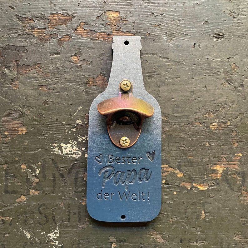 May include: A blue wooden bottle opener shaped like a beer bottle with a copper bottle opener. The bottle opener has the text "Bester Papa der Welt!" engraved on it.