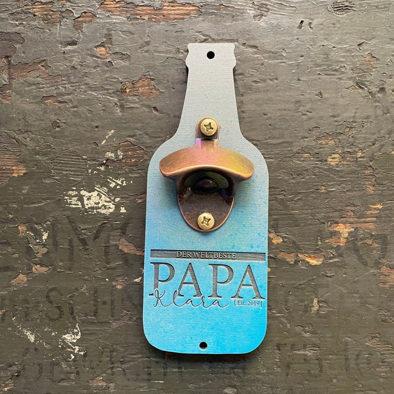 May include: A blue wooden bottle opener with a brass bottle opener attached. The bottle opener has the text "Der Weltbeste Papa Clara Est. 2019" engraved on it.
