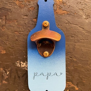 May include: A blue and white ombre bottle opener with a brass bottle opener attached. The bottle opener has the word "papa" written on it.