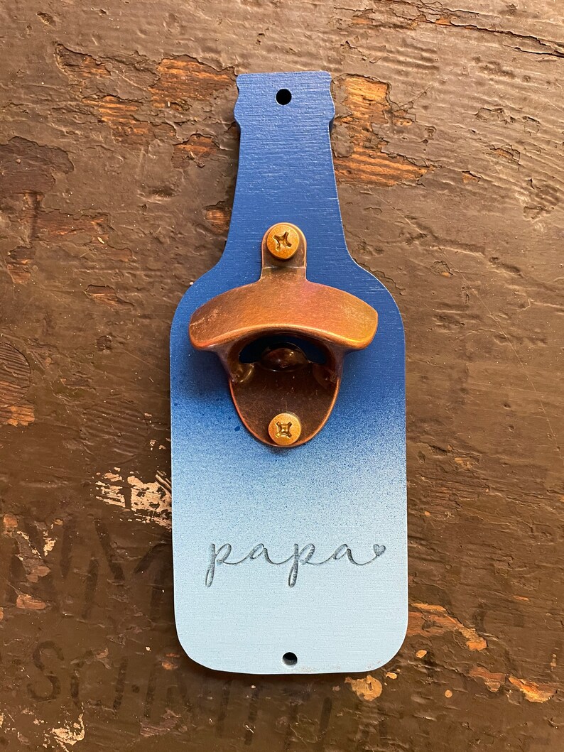 May include: A blue and white ombre bottle opener with a brass bottle opener attached. The bottle opener has the word "papa" written on it.