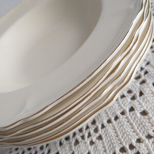 May include: A stack of cream-coloured ceramic bowls with a wavy edge and gold trim. The bowls are stacked, highlighting the design. The background is a white, open-weave fabric.