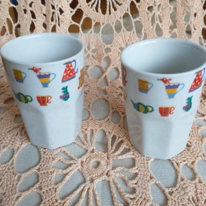 May include: Two white ceramic mugs with colourful illustrations of teacups, teapots, and other tea-related items. The mugs are sitting on a crocheted doily.