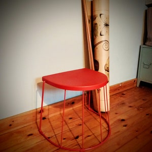 May include: A red metal stool with a curved, half-circle seat and a wire frame. The stool is sitting on a wooden floor.