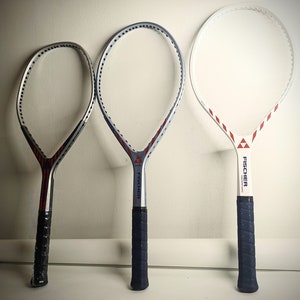 Op de afbeelding: Drie witte tennisrackets met blauwe en rode accenten. De rackets zijn allemaal van het merk "Fischer" en hebben een blauwe grip.