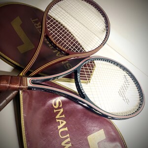 May include: Two vintage tennis rackets with brown and black handles and white stringing. The rackets are resting on top of two burgundy tennis racket bags with gold lettering that reads "Snauwaert".