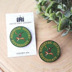 May include: Two circular wooden pins featuring a green background with a graphic of a person riding a creature while blowing a horn. The text reads MEDIEVAL HISTORY ENTHUSIAST in gold lettering. One pin is attached to a white card labelled CASTLE CREW COMPANY.