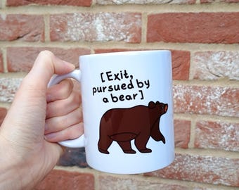 May include: White ceramic mug with the text "[Exit, pursued by a bear]" and a brown bear illustration. The mug has a curved handle and is held against a brick wall background.