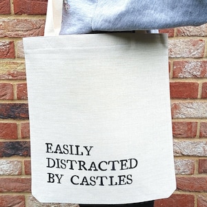 May include: A cream-coloured tote bag with the black text "EASILY DISTRACTED BY CASTLES". The bag is made of a textured fabric. The bag is being held in front of a red brick wall.