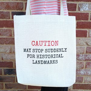May include: A cream-coloured tote bag with two shoulder straps. The front features the text CAUTION in red, followed by the black text MAY STOP SUDDENLY FOR HISTORICAL LANDMARKS. The bag has a visible woven texture and is made of a light-coloured fabric.