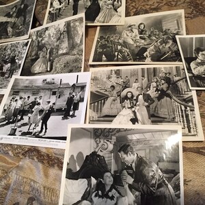 May include: A collection of black and white photographs from a film set. The photos show actors and actresses in period costumes, including a woman in a large hoop skirt and a man in a suit. The photos are arranged on a patterned surface.