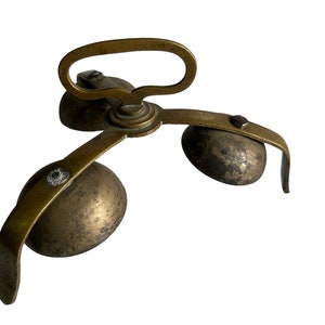 Puede incluir: Campana antigua de latón con un asa y tres campanas esféricas. La campana tiene un aspecto deslustrado y envejecido, lo que sugiere que es vintage. El asa es curva y las campanas están unidas a un mecanismo central.