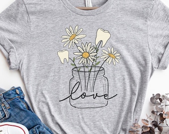 Op de afbeelding: Een grijs T-shirt met een afbeelding van een glazen pot gevuld met witte madeliefjes en tanden. De tekst "love" staat onder de pot.