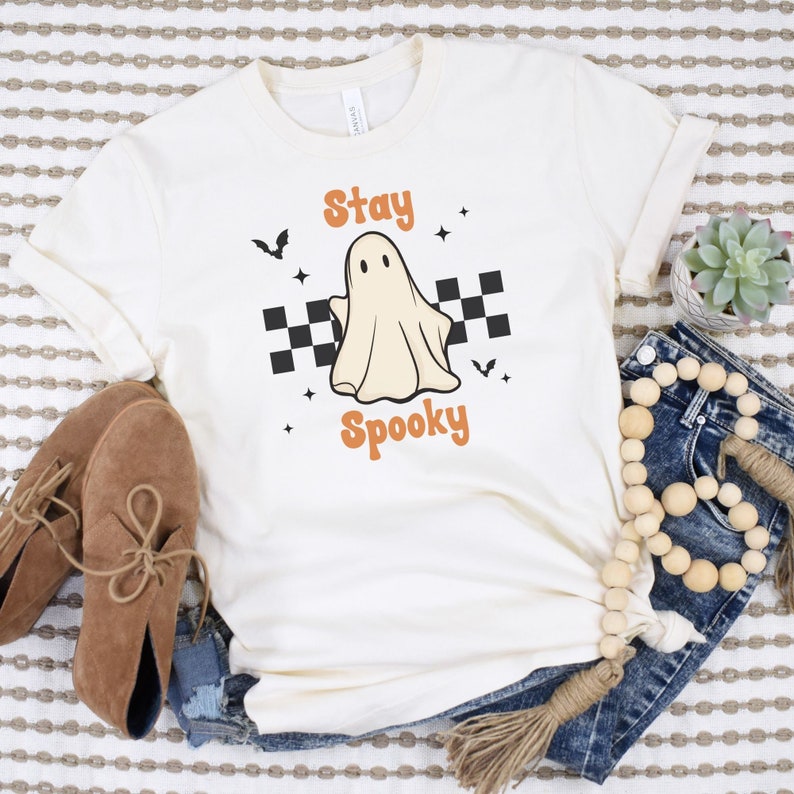 May include: Cream-colored t-shirt with the words "Stay Spooky" in orange, featuring a cartoon ghost and bats. The ghost is centered on a black and white checkered background. Stars and bats are scattered around the design.