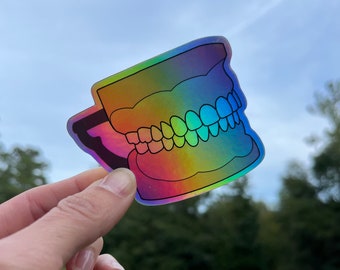Może przedstawiać: Holograficzna naklejka z zestawem zębów. Naklejka jest tęczowa i ma błyszczące wykończenie.