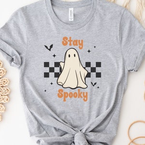 May include: A light gray t-shirt featuring a Halloween-themed design. The design includes a friendly ghost, the words "Stay Spooky" in orange, black bats, and star accents. A black and white checkered pattern is below the ghost.