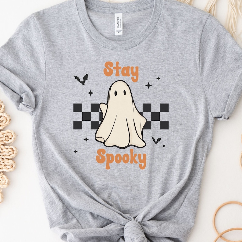 May include: A light gray t-shirt featuring a Halloween-themed design. The design includes a friendly ghost, the words "Stay Spooky" in orange, black bats, and star accents. A black and white checkered pattern is below the ghost.
