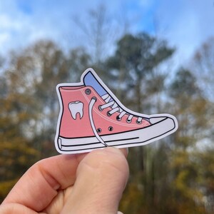 May include: Pink high-top sneaker with a white sole and a white tooth graphic on the side. The laces are white and the shoe is untied.