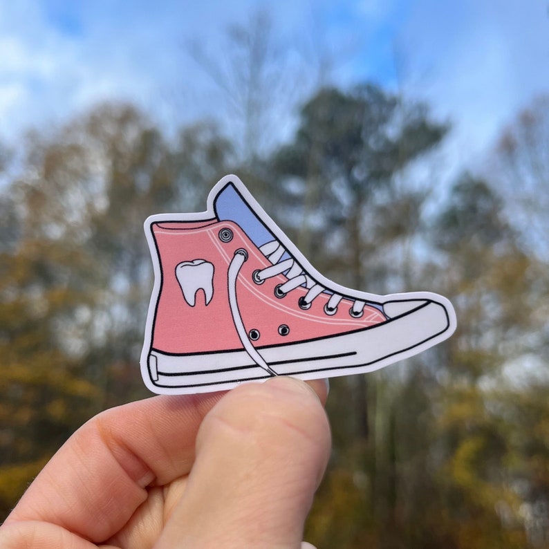 May include: Pink high-top sneaker with a white sole and a white tooth graphic on the side. The laces are white and the shoe is untied.