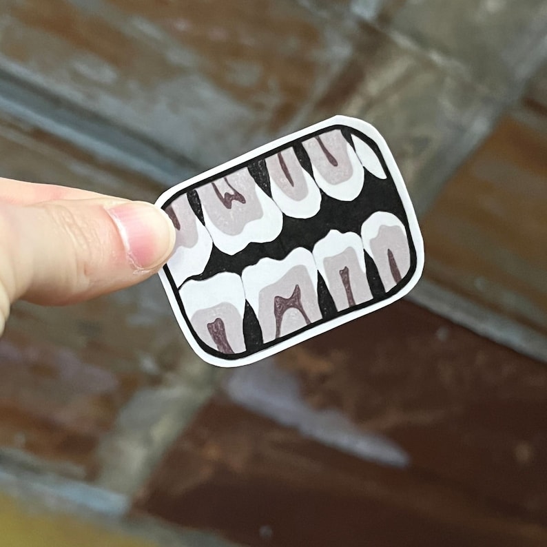 Op de afbeelding: Een close-up van een sticker met een set tanden. De tanden zijn wit met een grijze omtrek en zijn in een rij gerangschikt. De sticker is op een bruine achtergrond.