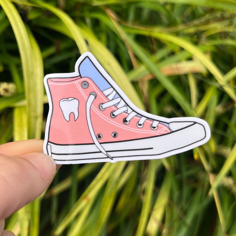 May include: Pink high-top sneaker with a white sole and white laces. The sneaker has a blue tongue and a white tooth graphic on the side.