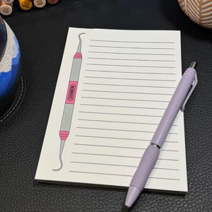 May include: A white notepad with horizontal lines, a light purple pen, and a dental tool illustration. The dental tool is gray with pink accents and the name "Shannon" printed on it. The pen is lying on the notepad.